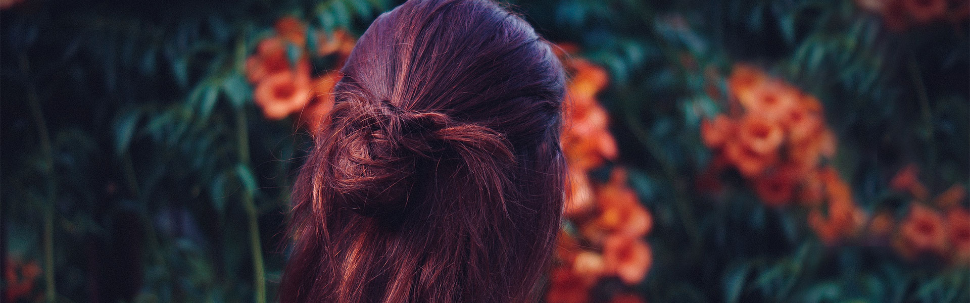 Frau mit langen roten Haaren von hinten vor einer Blumenhecke